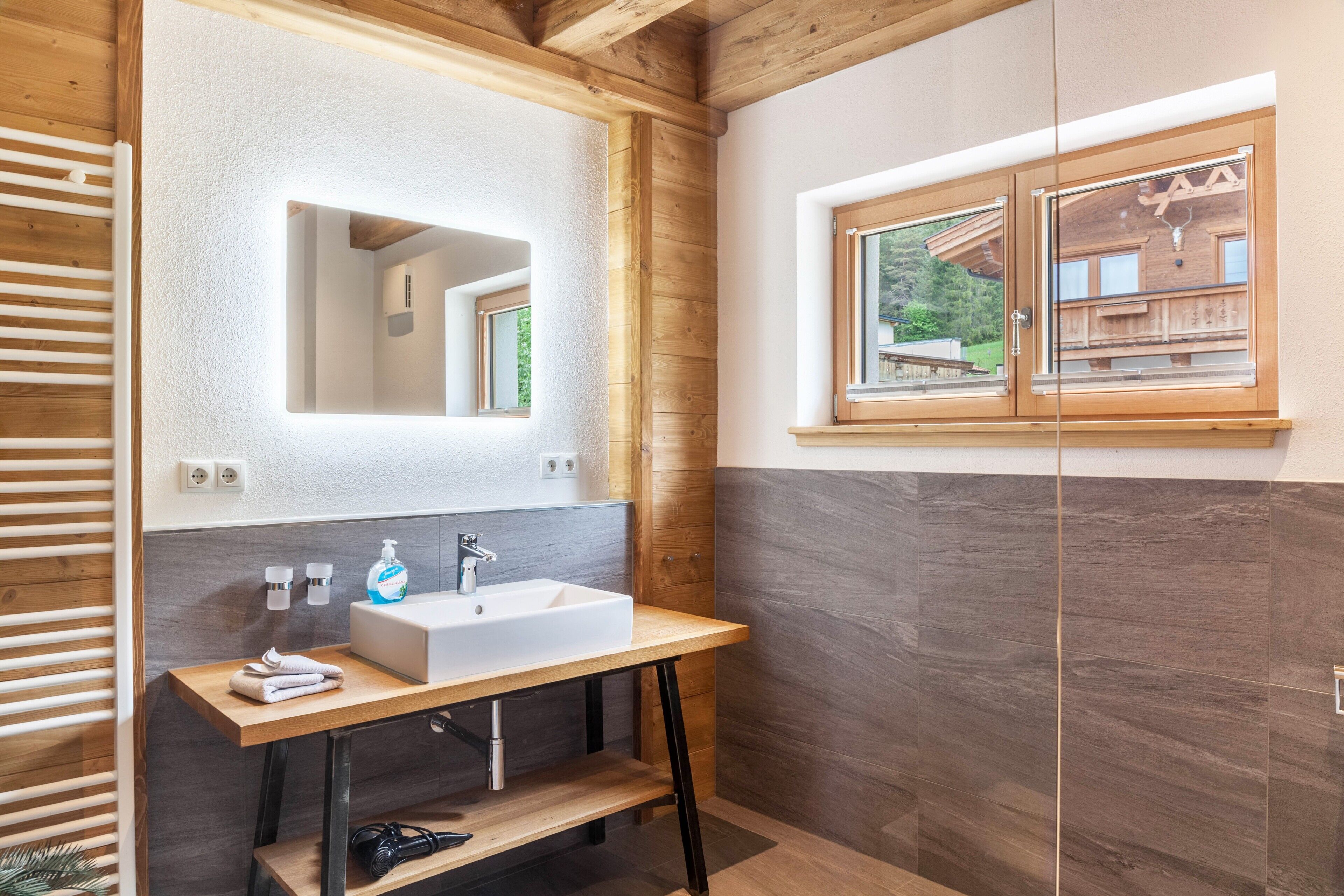 Ein modernes Badezimmer mit einem Holzwaschtisch, einem großen Spiegel, einem Fenster mit Blick auf eine Holzhütte und einem Handtuchhalter.