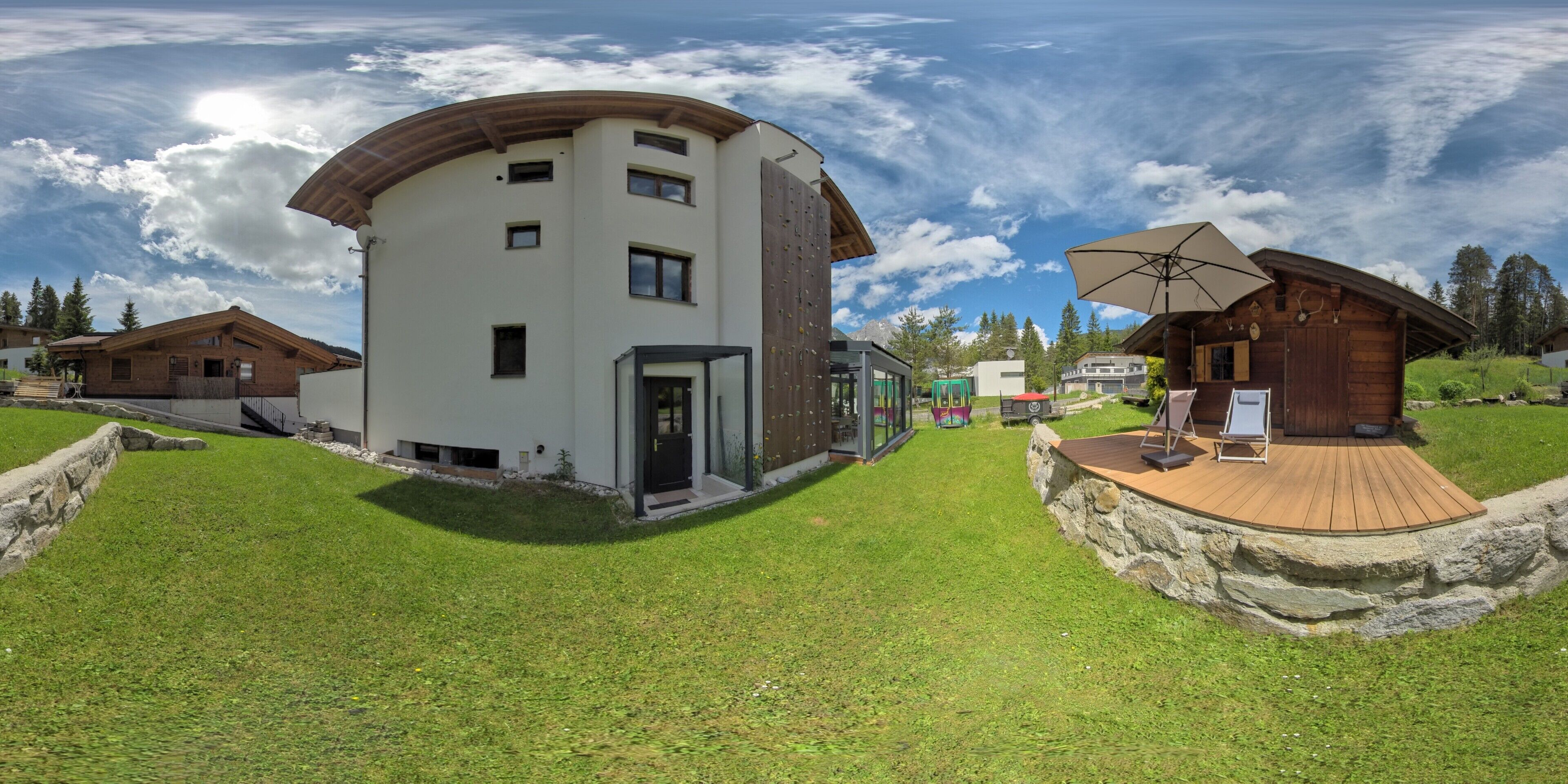 Ein modernes, mehrgeschossiges Gebäude mit gewölbter Dachform, einer Holzdiele mit Tisch und Stühlen sowie einer glasgeschlossenen Struktur.