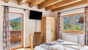 A bedroom with a wooden ceiling, a bed with pillows, a wooden wardrobe, a television, and a window with a view of mountains.