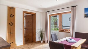 Ein Zimmer mit Holztür, einem Tisch mit rotem Tischläufer, einem Stuhl, einer Topfpflanze und Blick auf einen Balkon.