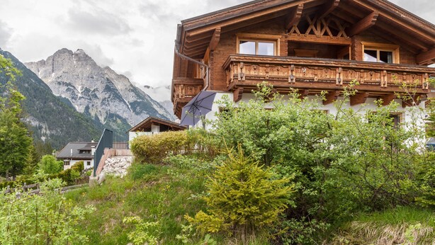 Eine Holzchalet mit Balkon, umgeben von üppiger Vegetation und Bergen im Hintergrund.