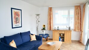 A living room with a blue sofa, a wooden coffee table, a small table with a vase and sunflower, and a kitchen area with a wooden cabinet and window with curtains.