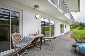 A covered outdoor patio with a table and chairs, a clock, and a view of the greenery.