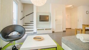 Un salon moderne avec un téléviseur à écran plat, un fauteuil noir, une table basse blanche, un canapé et un escalier menant à une autre pièce.