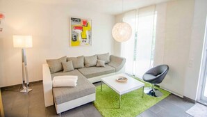 Un salon moderne avec un canapé d’angle, une table basse blanche, un fauteuil noir, une lampe sur pied et une grande fenêtre laissant entrer la lumière naturelle.