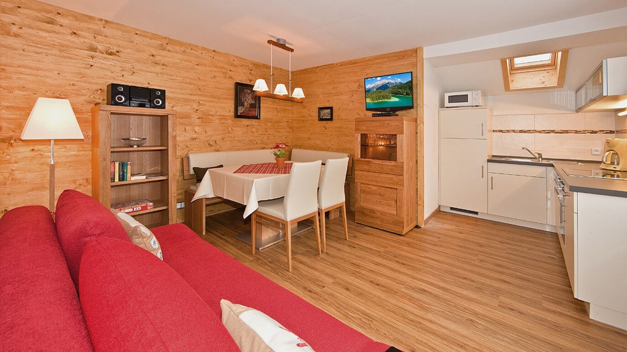 Une pièce chaleureuse avec un canapé rouge, une table à manger avec des chaises, un téléviseur, un coin cuisine et des murs en bois.