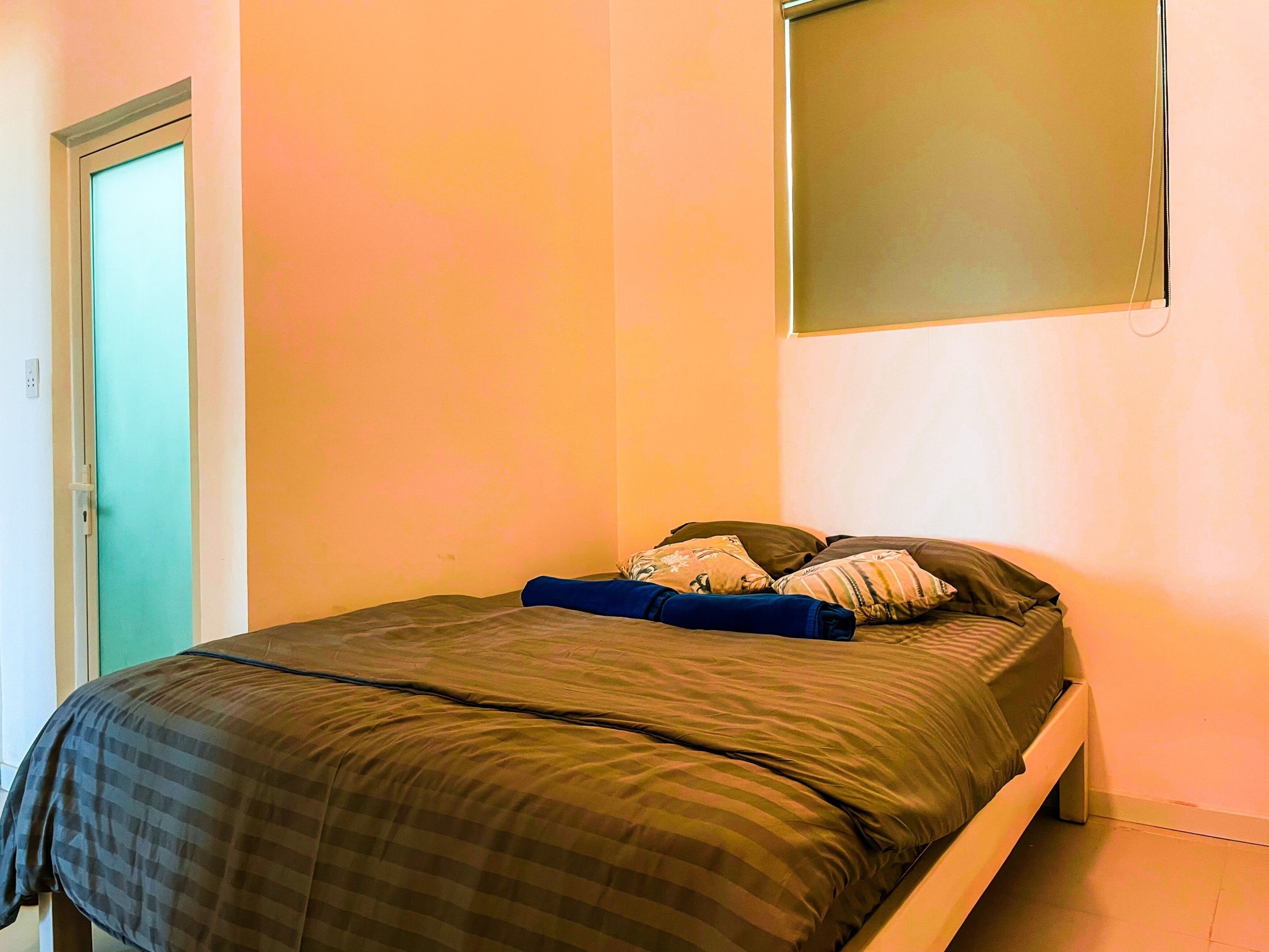 A single bed with a dark checkered bedspread and multiple pillows, a plain wall, and a door with a light switch.