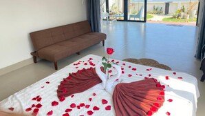 A bedroom with a bed adorned with rose petals and a swan decoration, a brown sofa, and a view of the ocean through large windows. - Jeju Beach Front Villa (Da Nang)