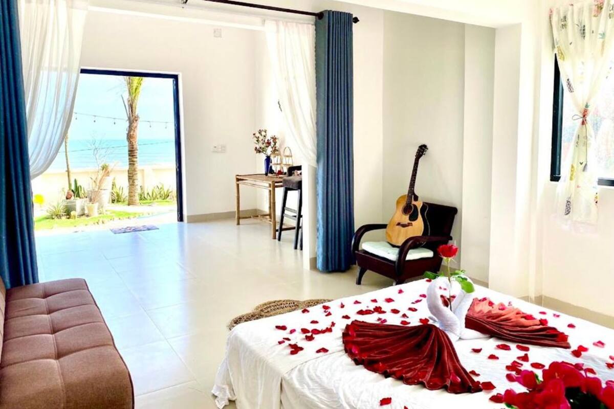 Un dormitorio con una cama cubierta de pétalos de rosa, una guitarra y vista al mar a través de un gran ventanal.