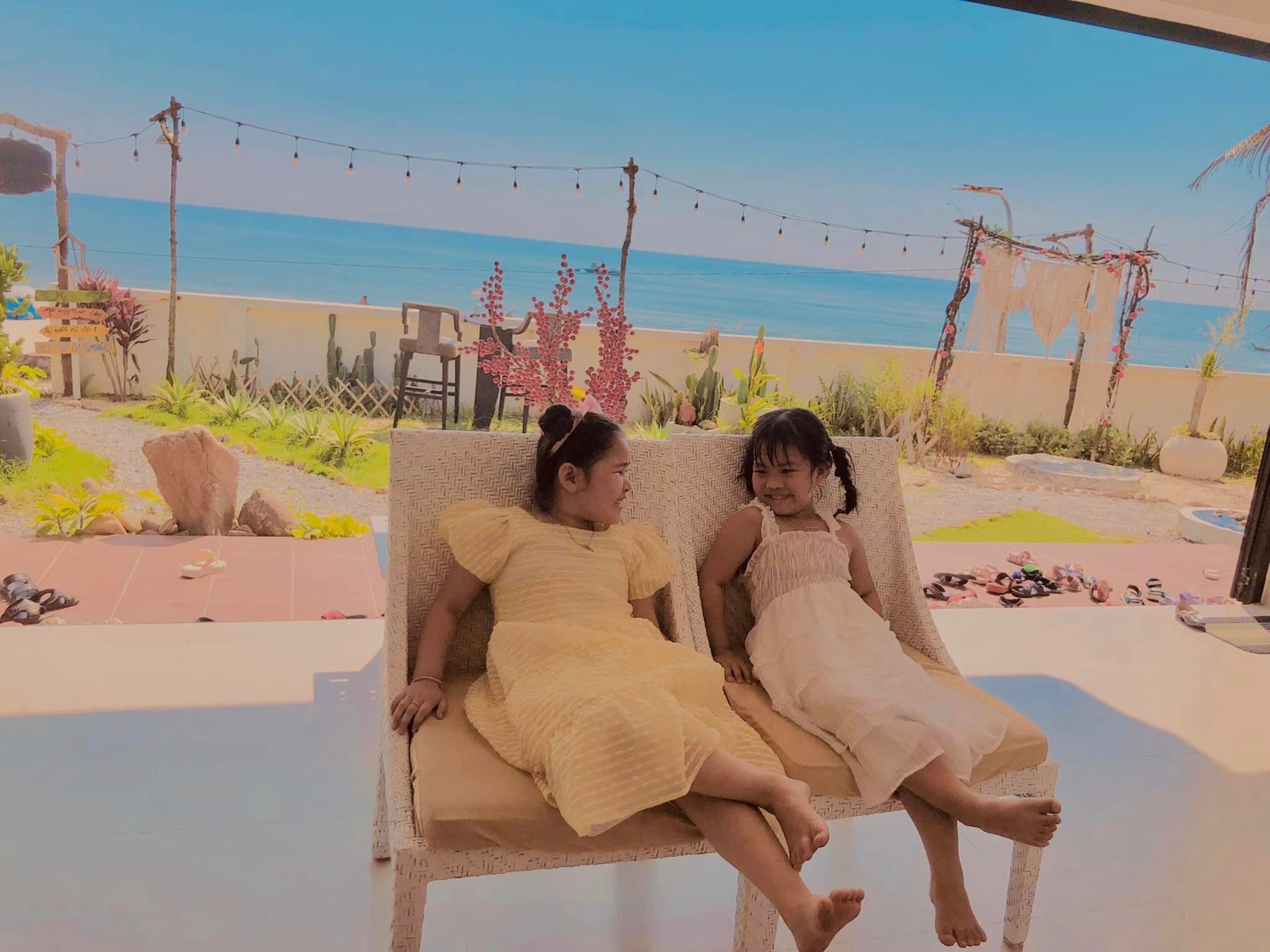 Deux enfants sont assis sur un banc en osier, à l’extérieur, avec vue sur la plage et des guirlandes lumineuses en arrière-plan.
