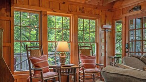 A wooden cabin interior with large windows, two rocking chairs, a table with a lamp, and a couch.