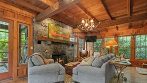 A cozy cabin interior with a stone fireplace, two grey sofas, a wooden ceiling, and large windows offering a view of the forest.