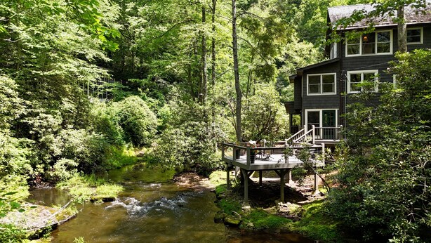 A cabin with a deck overlooking a forested area and a stream.