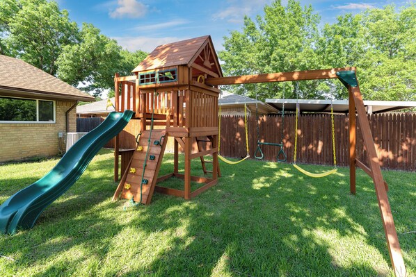 A wooden play structure with a slide and climbing area, set in a backyard with a wooden fence and trees in the background.