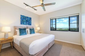 Ein Schlafzimmer mit einem Bett, zwei Nachttischen, einem Deckenventilator, einem großen Fenster und einem blauen Blumenbild.