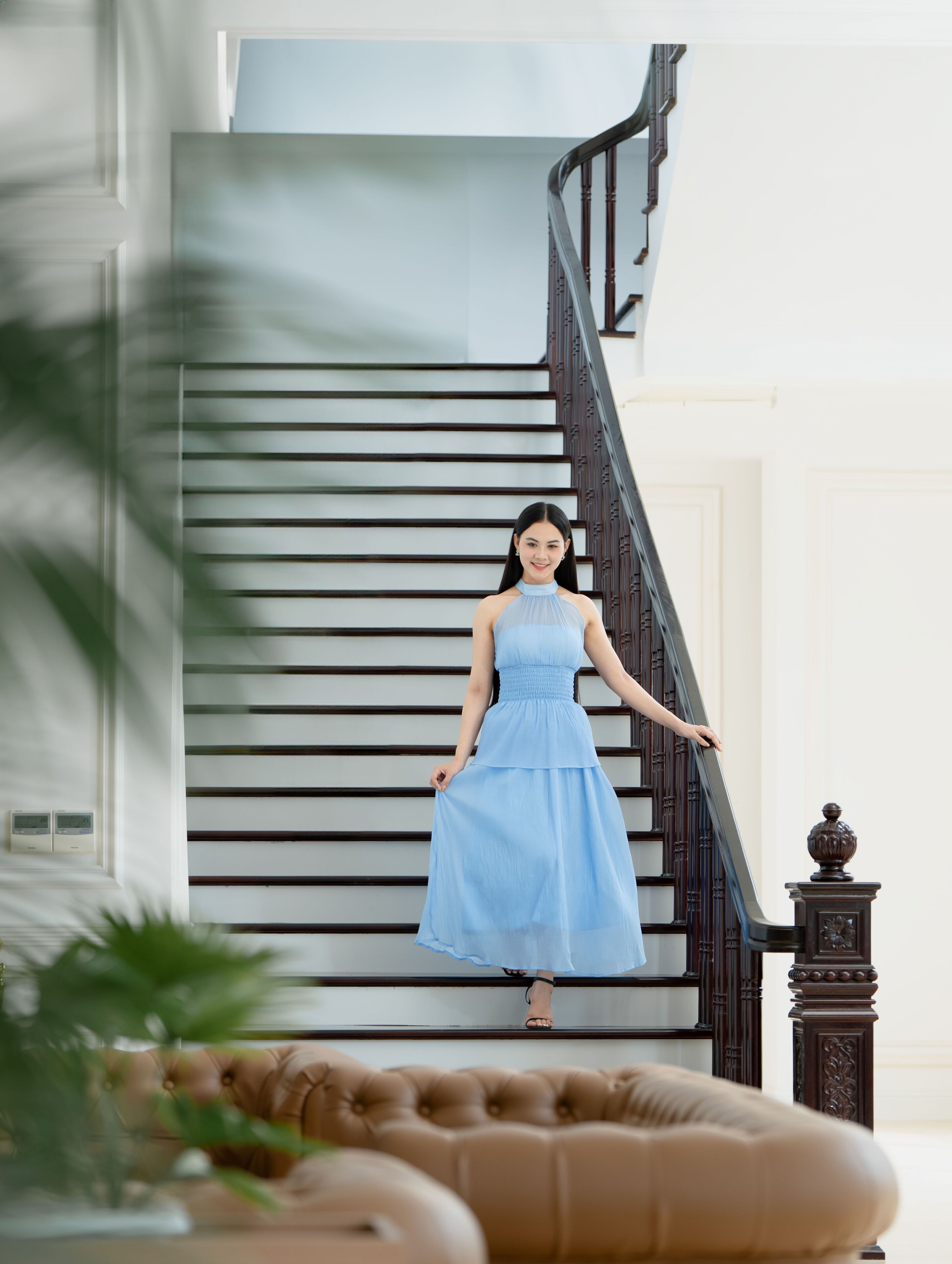 Une femme vêtue d’une robe bleu clair, debout sur un escalier dont les rampes sont en bois foncé, avec un mur blanc en arrière-plan.