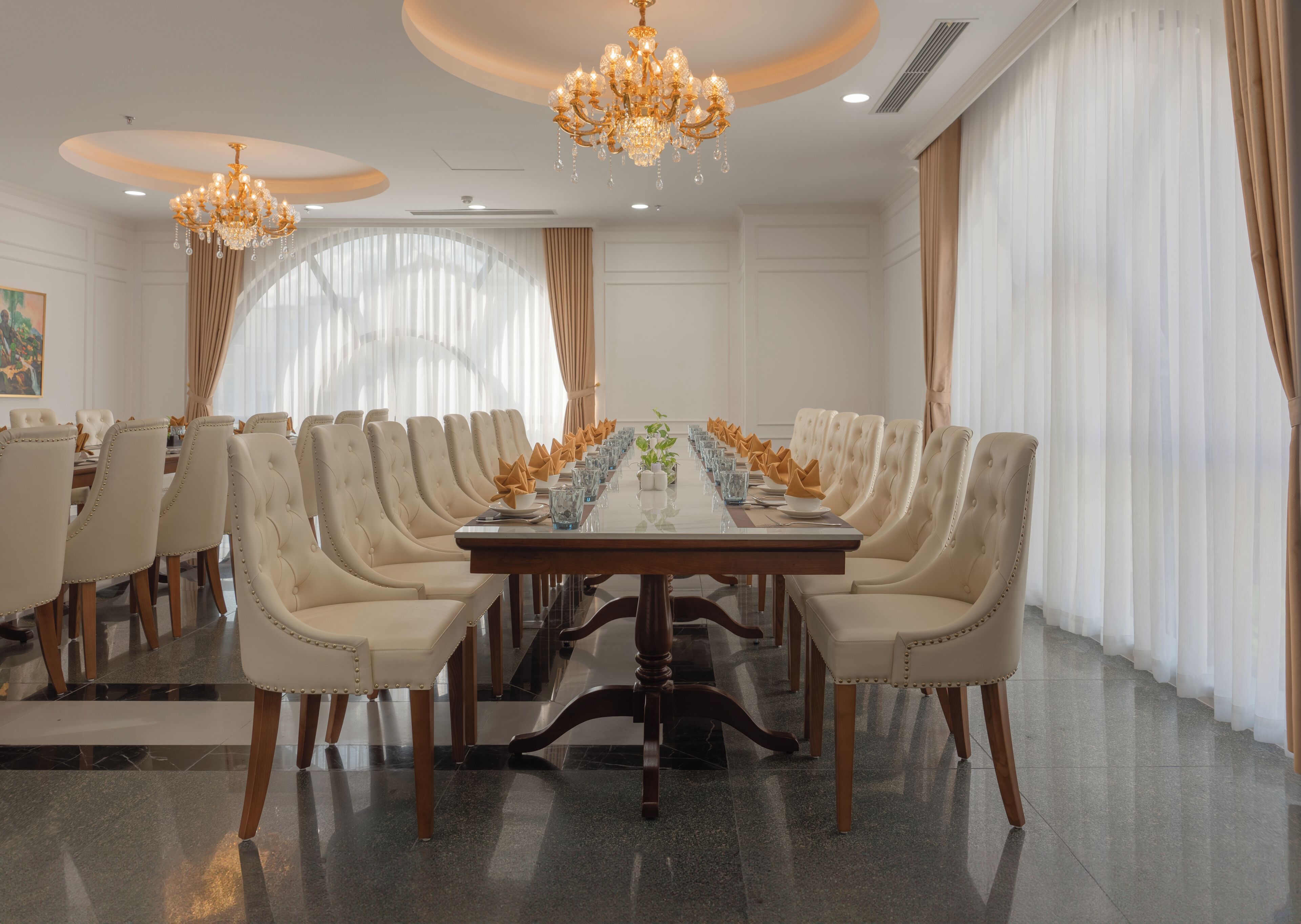 A formal dining room with a long table, multiple chairs, chandeliers, and large windows with curtains.