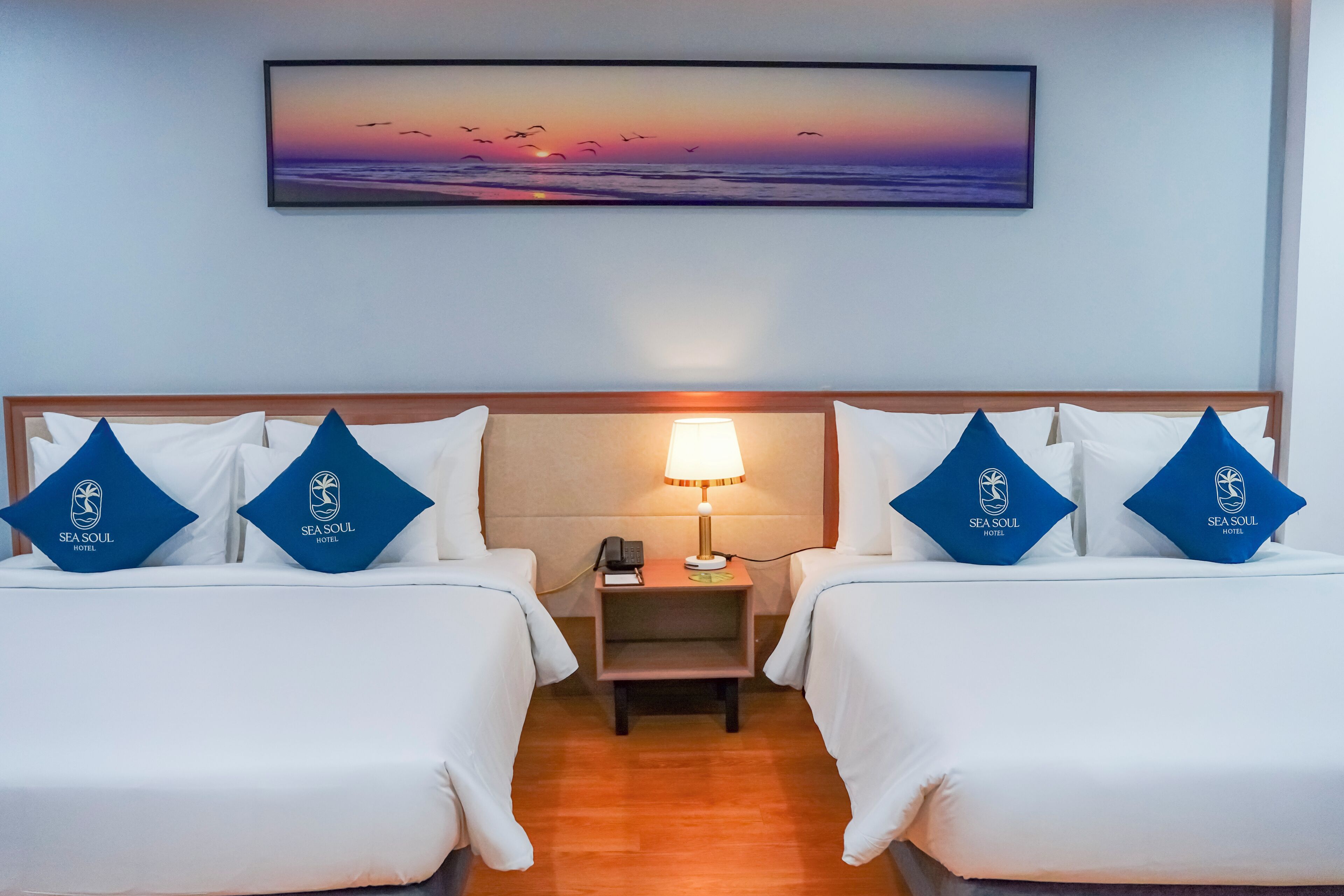 Une chambre d’hôtel avec deux lits, une table de chevet avec une lampe, un tableau représentant un coucher de soleil et des oreillers ornés du logo de l’hôtel.