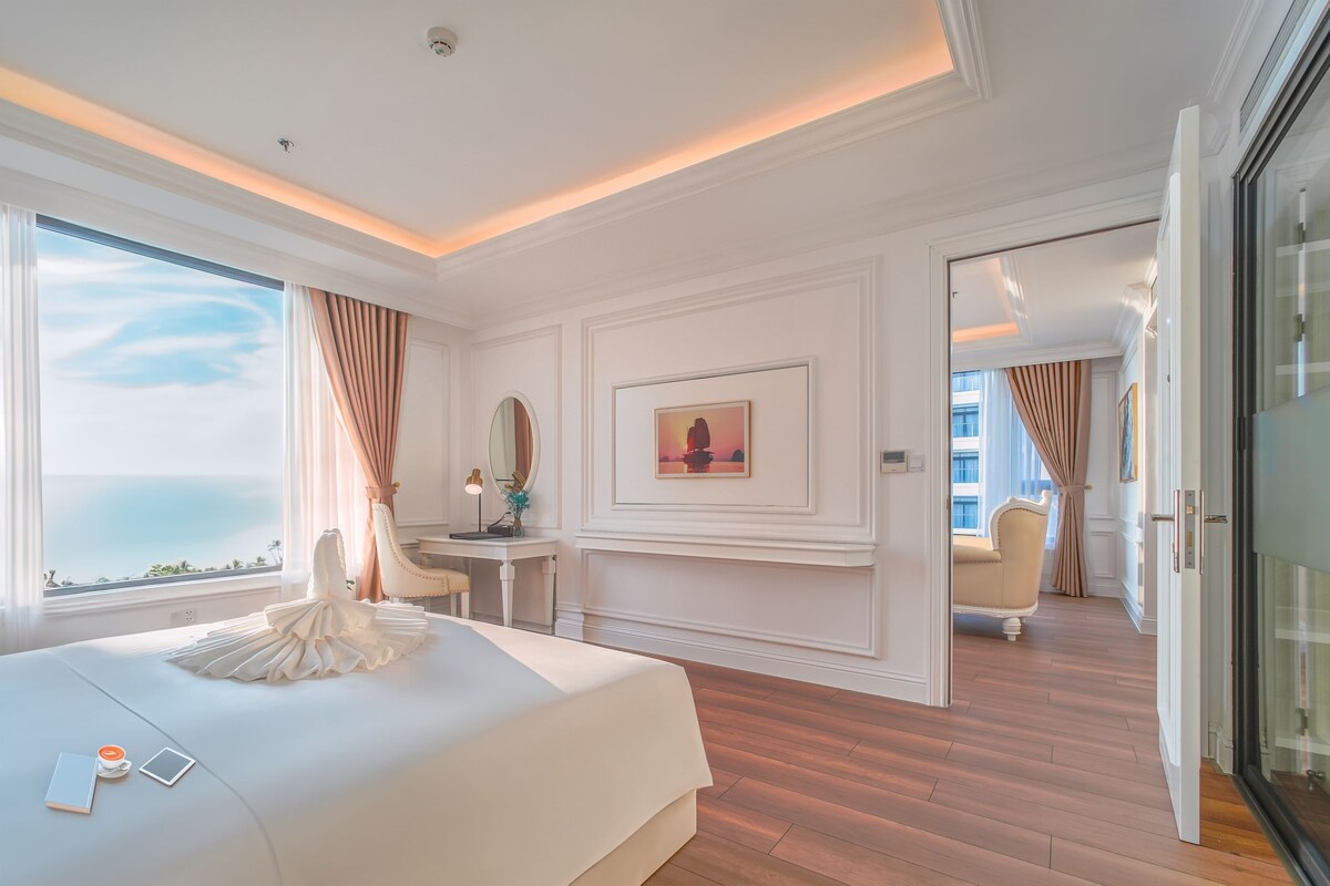 A hotel room with a bed, a desk, a chair, a large window with curtains, and a painting on the wall.