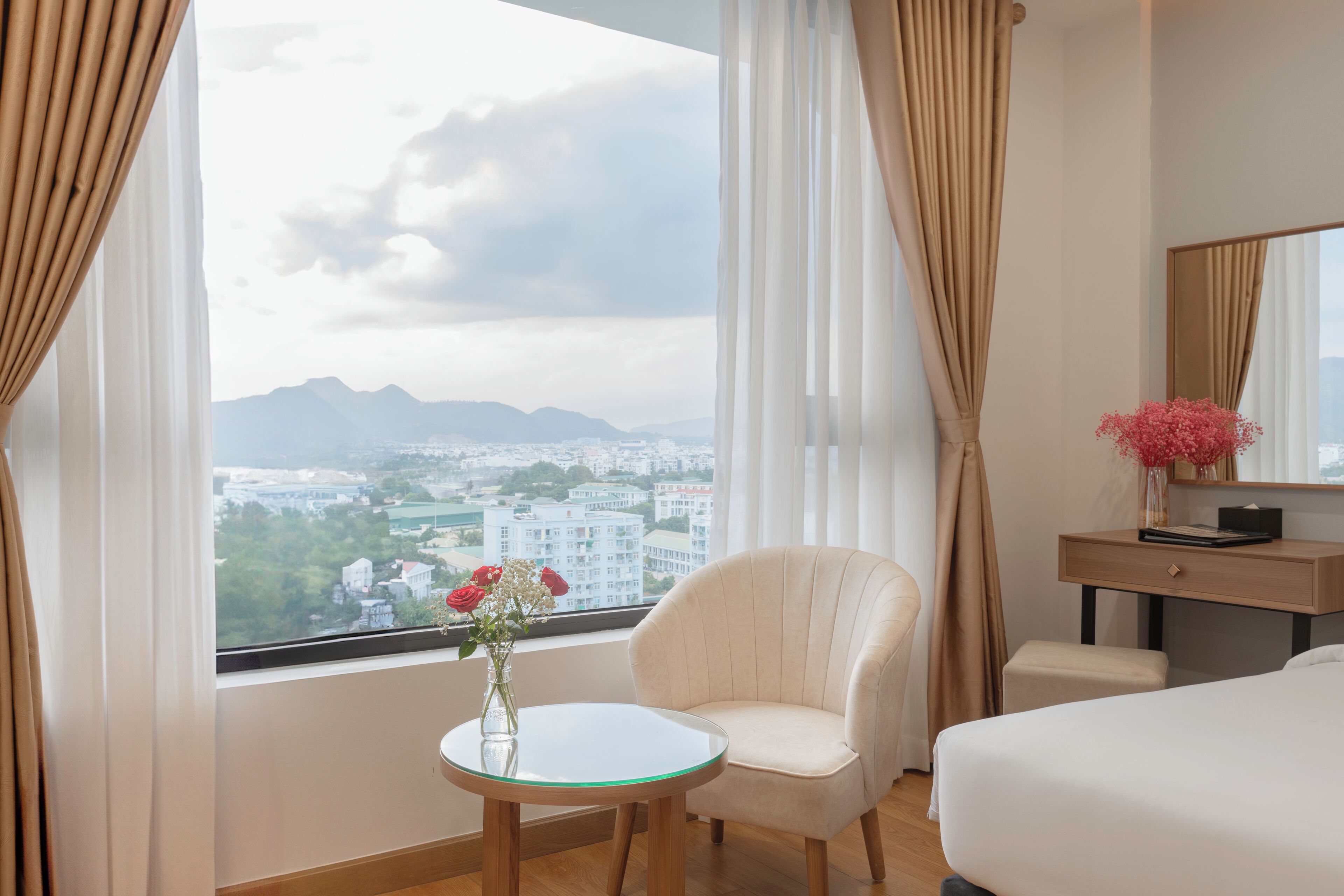 A hotel room with a bed, a chair, a small table with a vase of flowers, a desk, and a large window offering a city view.