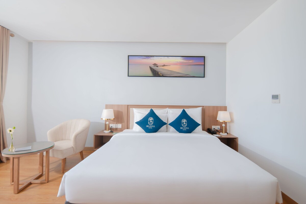 A hotel room with a bed, two pillows, a nightstand, a lamp, a chair, a small table, and a painting of a sunset over water.