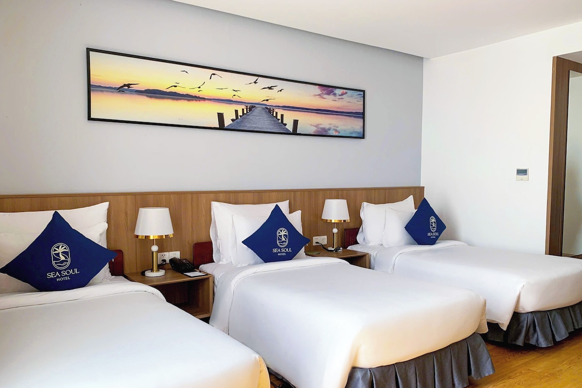 A hotel room with two beds, each with a pillow featuring the hotel's logo, a nightstand with a lamp, and a painting of a sunset over a pier.