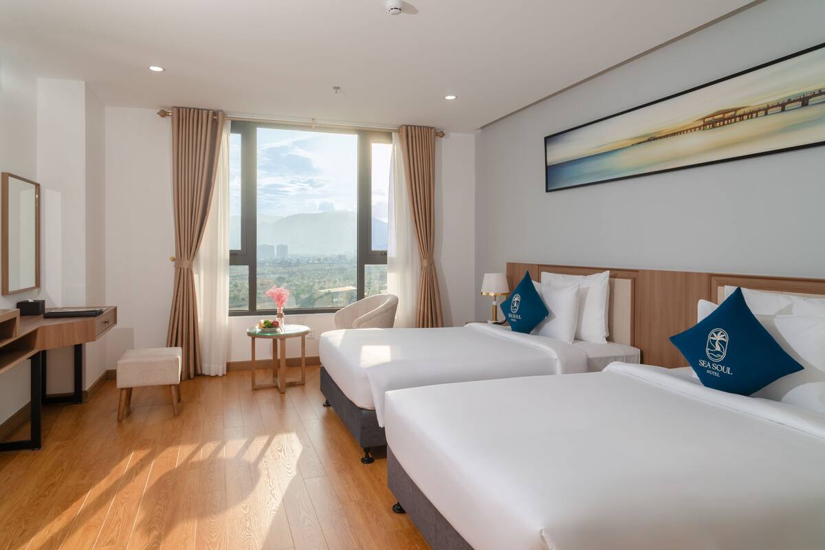 A hotel room with two beds, a desk, a chair, a small table, and a large window with curtains.