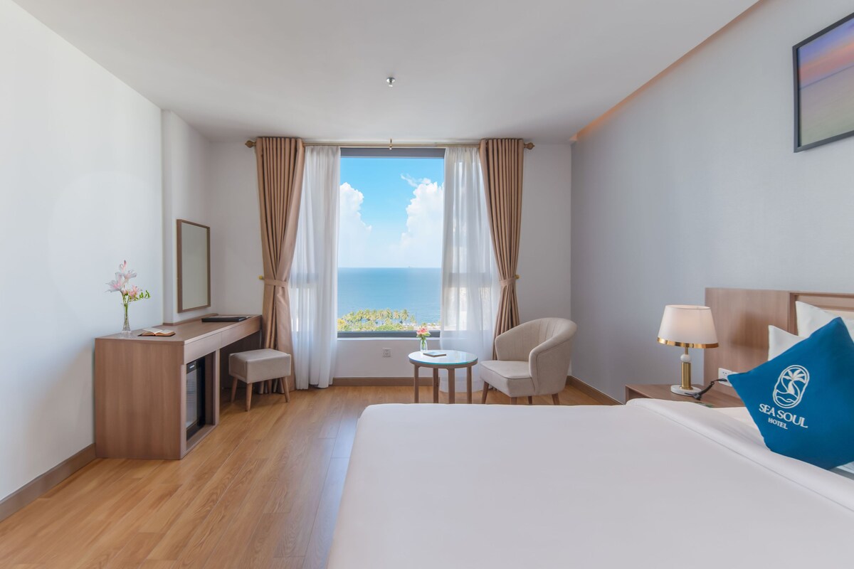 A hotel room with a bed, a desk, a chair, a small table, and a view of the sea.