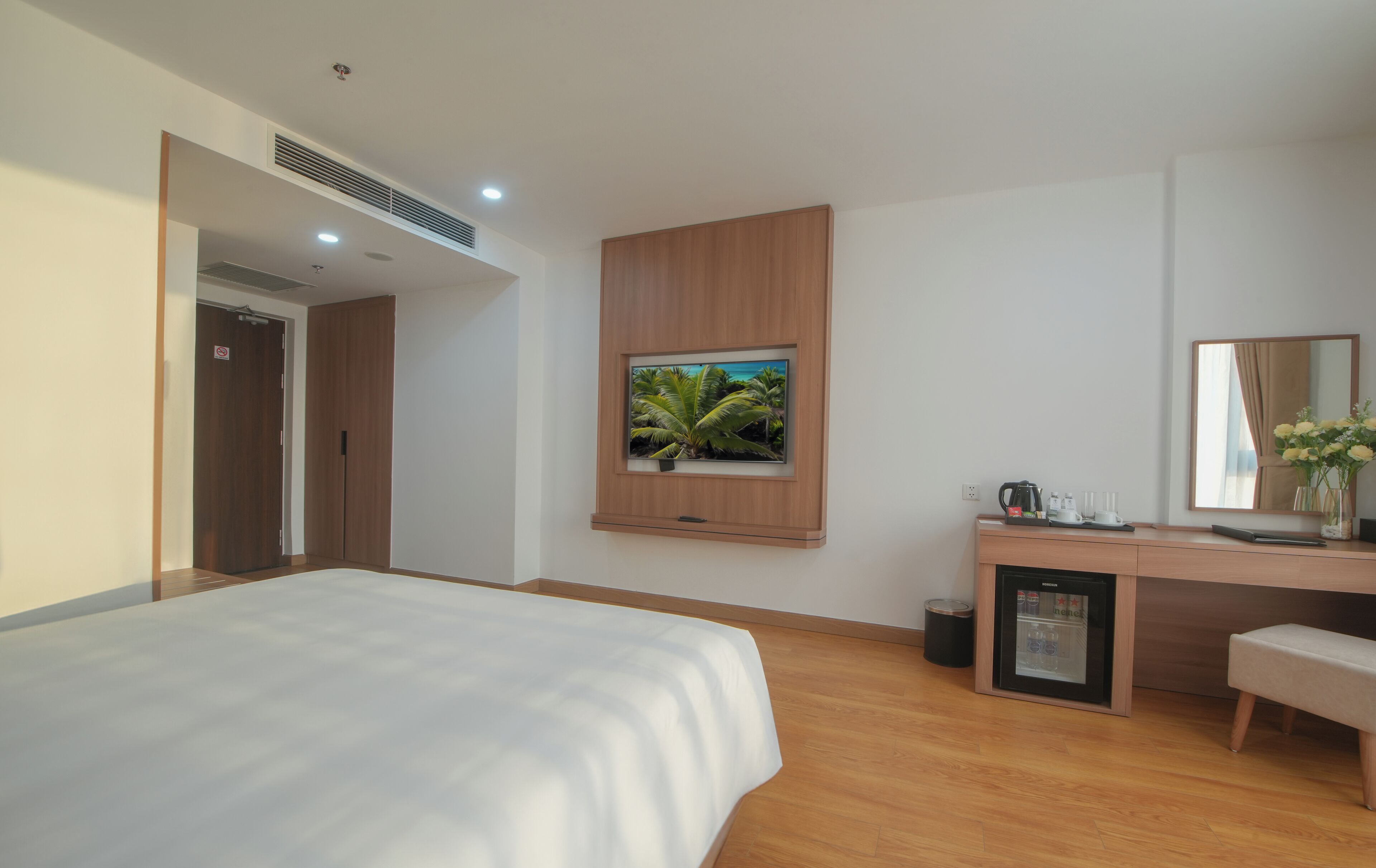 Una habitación de hotel moderna con una cama grande, un televisor montado, un escritorio con cafetera y un refrigerador pequeño.