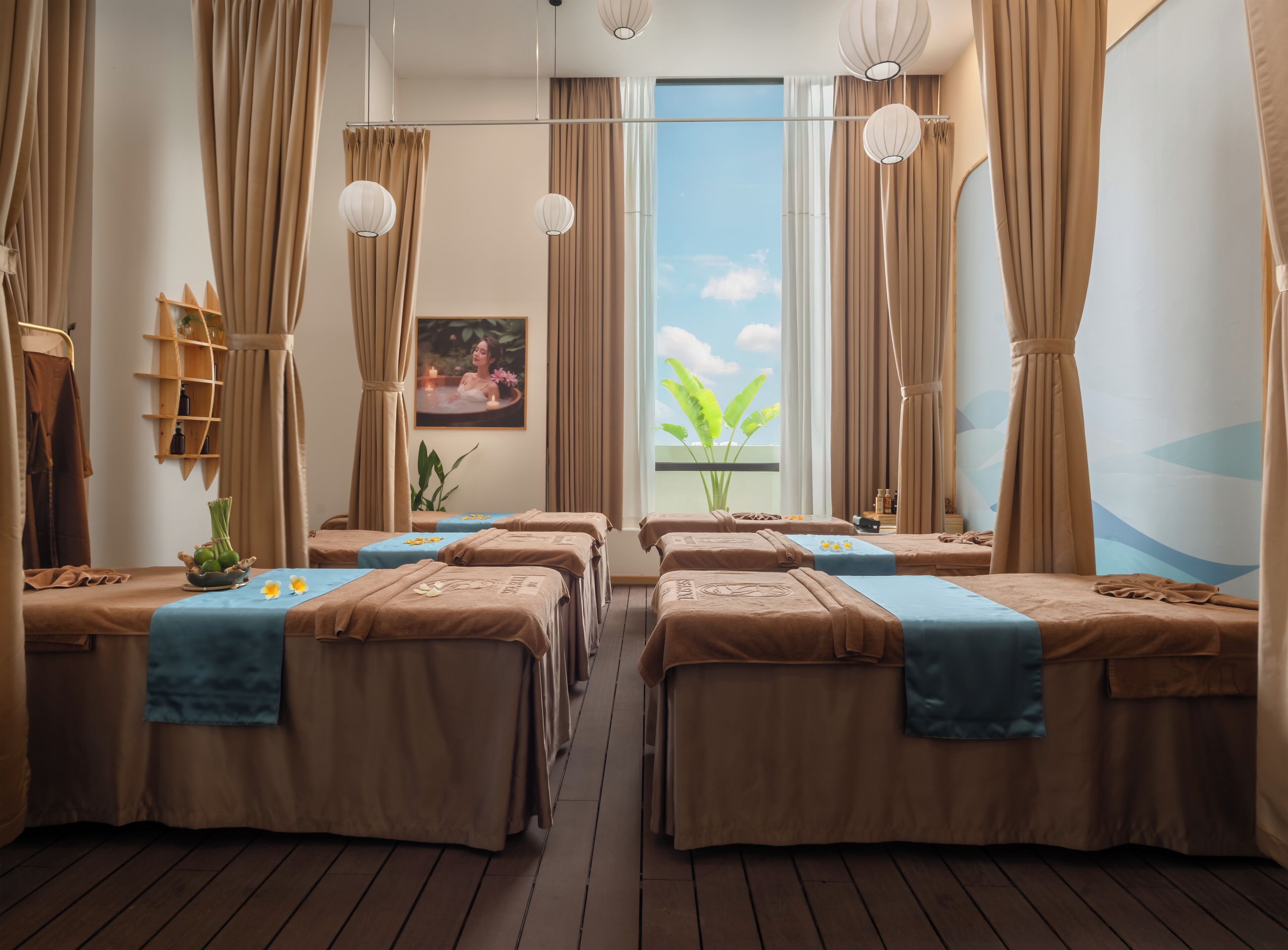 Une salle de spa équipée de quatre tables de massage, chacune agrémentée d’une serviette et d’une petite plante. On y trouve deux grandes fenêtres avec des rideaux, une étagère en bois où sont disposées des bouteilles, et un tableau accroché au mur.