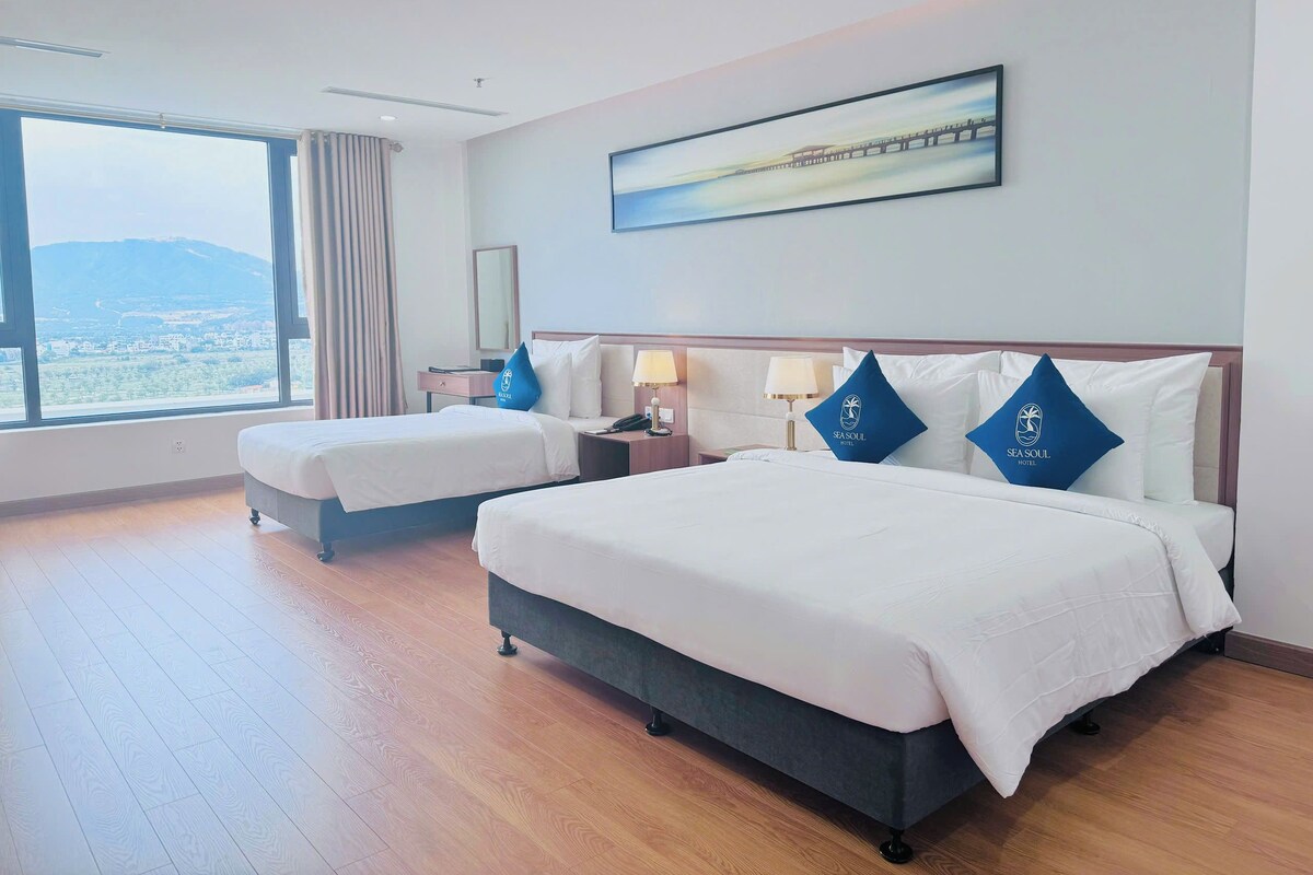 A hotel room with two beds, a large window with a view, a painting on the wall, and a nightstand with a lamp.