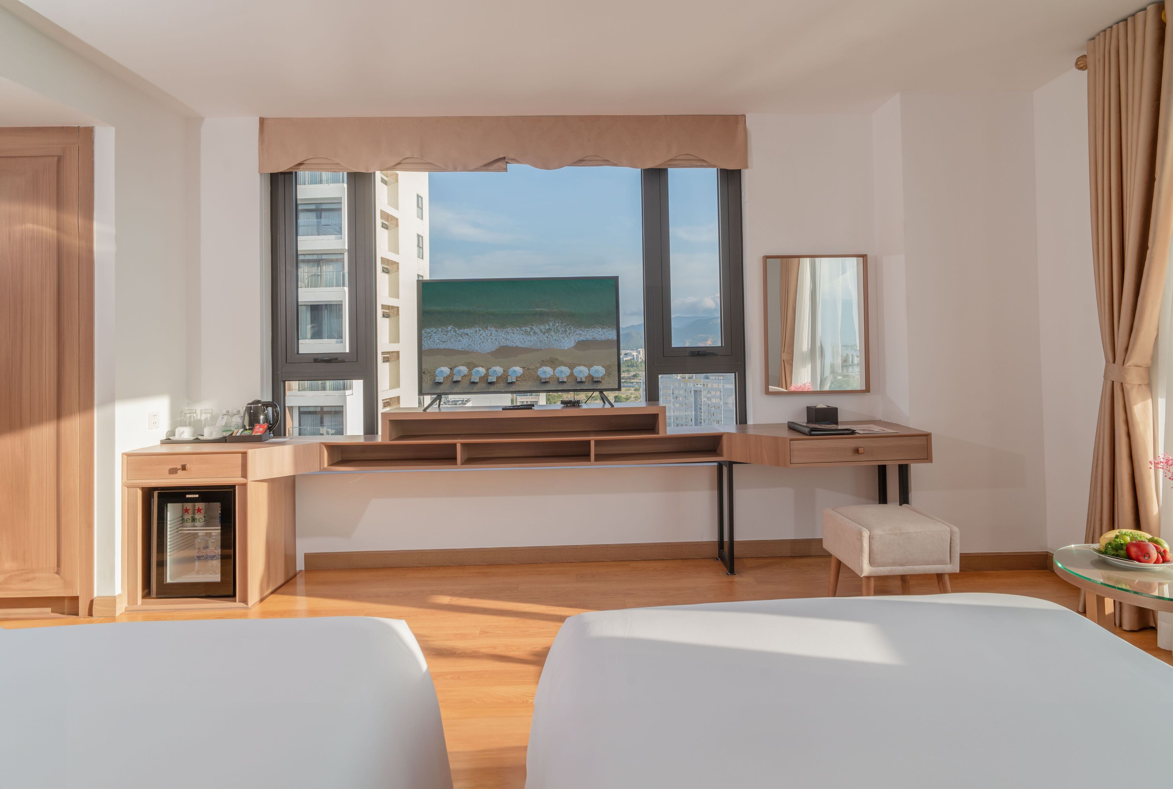 A hotel room with a large window, a desk with a computer, a refrigerator, and two beds.