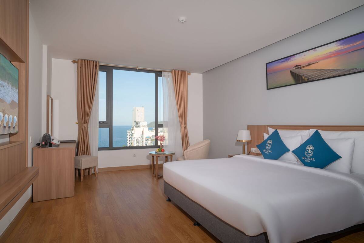 A hotel room with a large bed, a desk, a chair, a small table, and a large window with curtains.