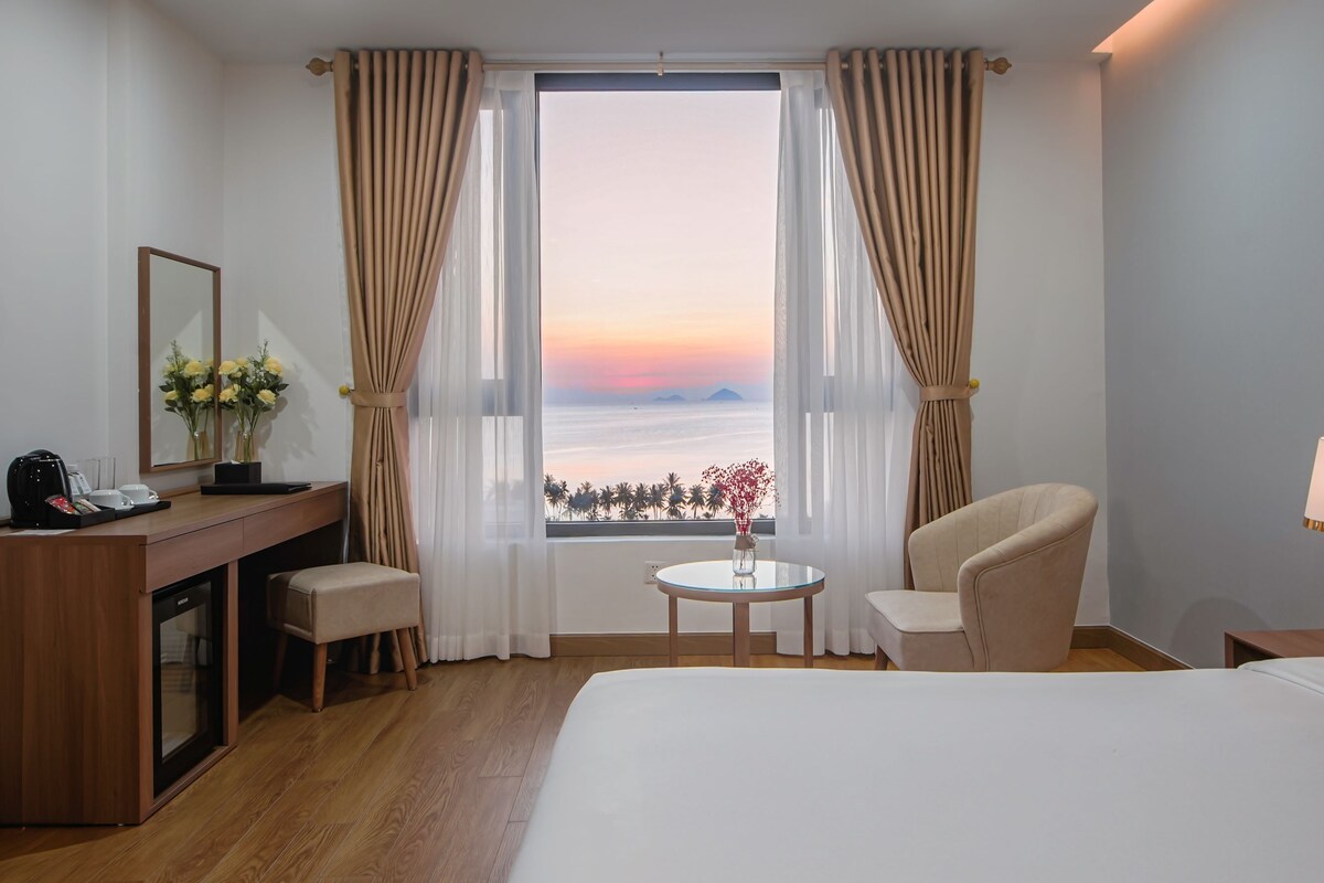 A hotel room with a bed, a desk with a phone and a lamp, a chair, a small table with flowers, and a window with curtains.