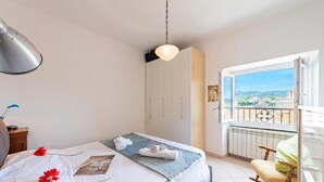 A bedroom with a bed, a bedside table, a chair, a window with a view, and a hanging light fixture. - Kimera Del Parasio Historic Apartment for 4 (Imperia)