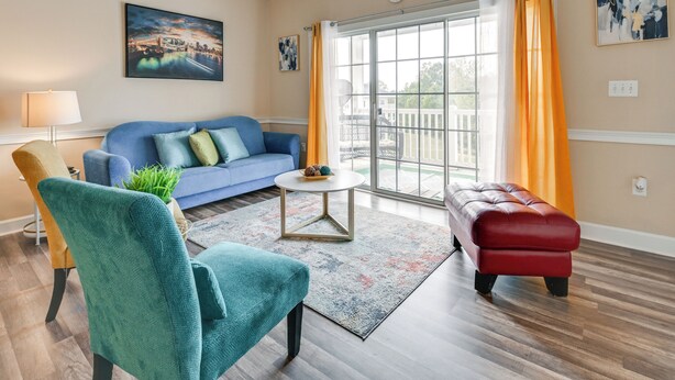 A living room with a blue sofa, teal armchair, and red ottoman, a round coffee table, and a large window with curtains.