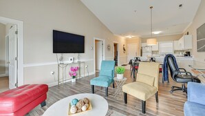 A spacious living area with a flat-screen TV, a red bench, a round coffee table with decorative items, and a kitchen in the background.
