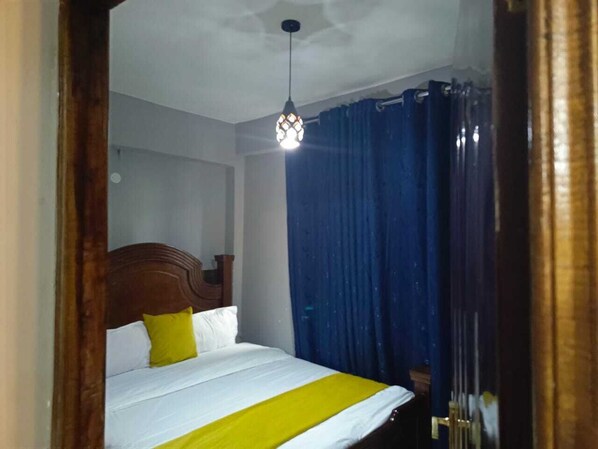 A bedroom with a bed, a wooden headboard, a yellow and white bedspread, a hanging light fixture, and blue curtains.
