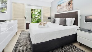 Un dormitorio moderno con cama, mesitas de noche, ventilador de techo y vistas a un jardín por un ventanal corredero.