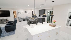 A modern living room with a flat-screen TV, a sofa, and a dining area with a white island and pendant lights. - Close to the Beach and 5th Ave! Fun Naples Peaceful Getaway 4U!  (Naples)