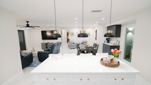 A modern kitchen with white cabinetry, a central island, pendant lighting, and a dining area with a sofa and table. - Close to the Beach and 5th Ave! Fun Naples Peaceful Getaway 4U!  (Naples)