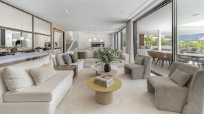 Un salon moderne avec un canapé, une table basse et des fauteuils, une cuisine en arrière-plan et de grandes fenêtres offrant une vue dégagée.