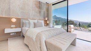 A modern bedroom with a large bed, bedside tables, a bench, and a view of a mountainous landscape. - Villa Almost Heaven by Vacation Marbella (Nueva Andalucía)