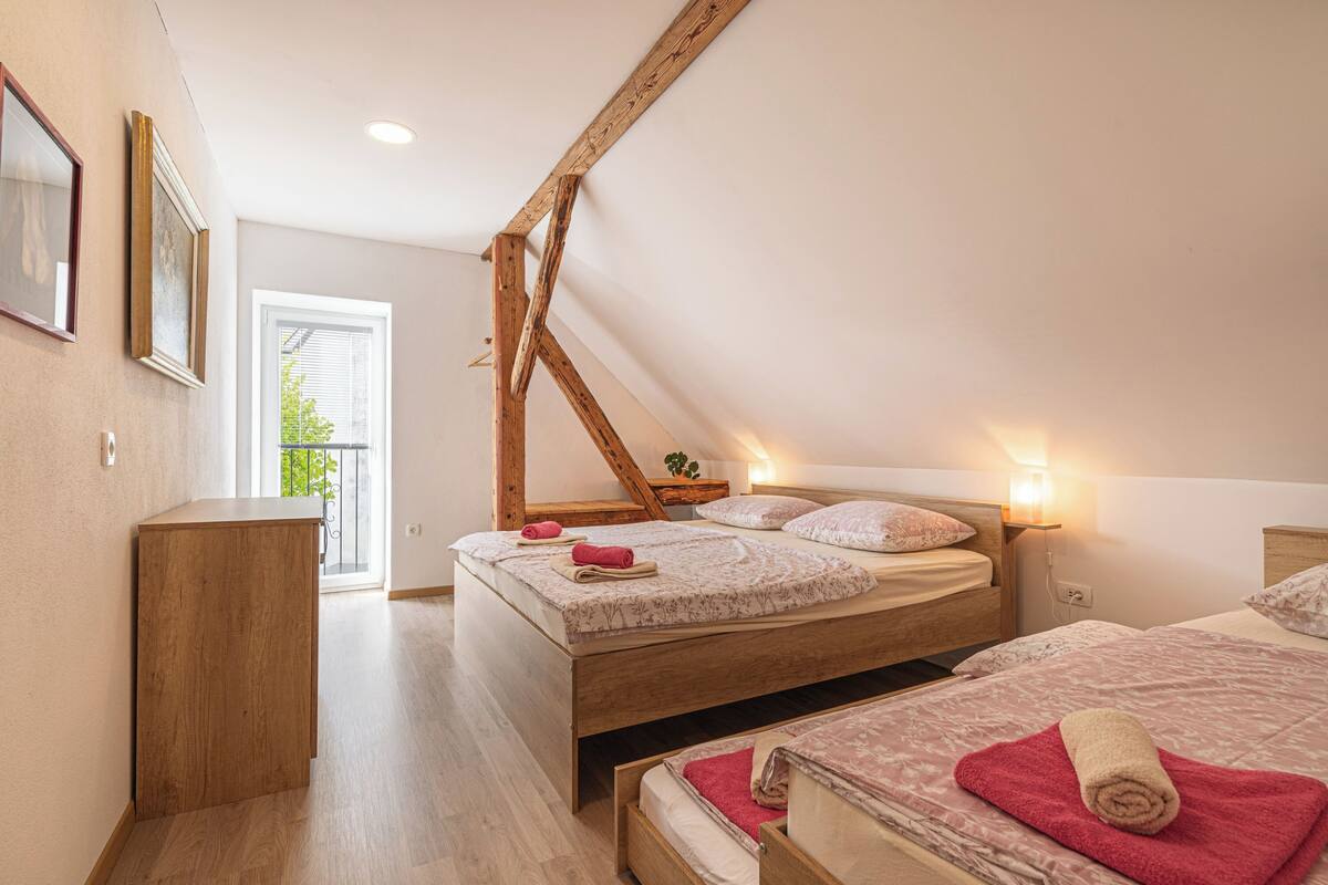 A bedroom with a sloped ceiling, wooden beams, two beds, a wooden cabinet, and a window with a view of greenery.