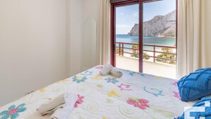 A hotel room with a bed, a window with a view of the sea and a mountain, and a balcony with a railing.