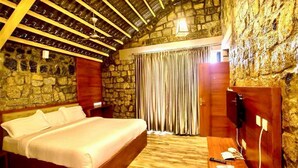 A bedroom with a stone wall, a wooden ceiling, a bed, a desk, and a TV. - Leopard Rock Wilderness Resort Ooty by VOYE HOMES (Ootacamund)