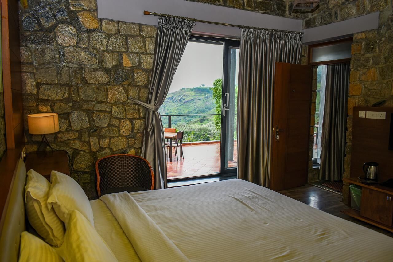 Une chambre avec un mur en pierre, un lit avec des oreillers, une chaise, une table et une vue sur un balcon agrémenté d’une chaise et de plantes.