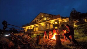 A modern house with a large glass facade, a fire pit in the front, and a group of people gathered around it. - Leopard Rock Wilderness Resort Ooty by VOYE HOMES (Ootacamund)