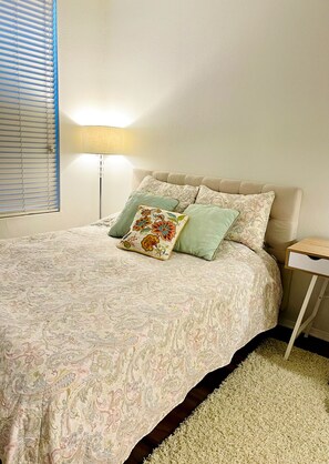 Une chambre à coucher avec un lit, des oreillers, une table de chevet, une lampe et une fenêtre avec des stores.