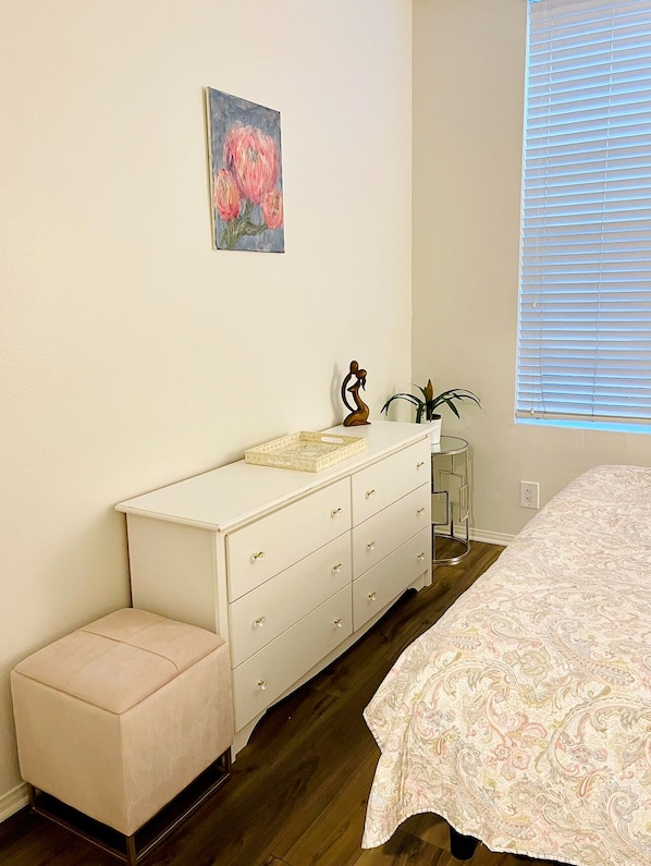 Un dormitorio con una cama, un tocador, un taburete pequeño, una planta en maceta y un cuadro en la pared.